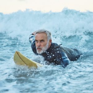Beach, surfer and old man on surfboard swimming in nature enjoying dangerous water sports and retir