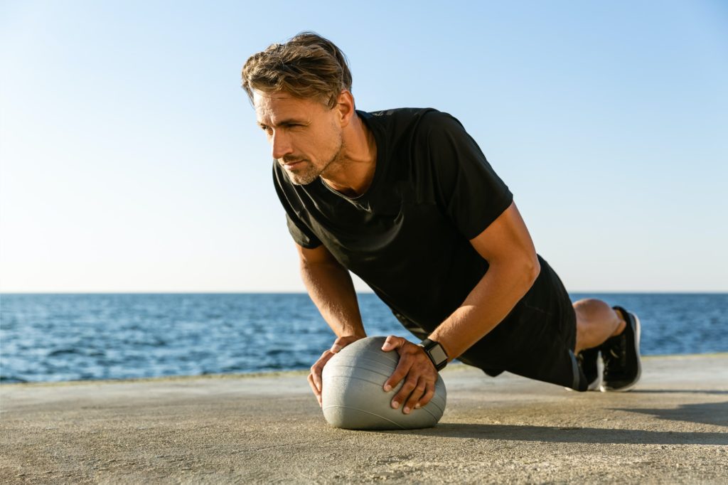 strong adult sportsman doing push ups with fit ball on seashore